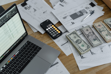 A professional office desk with financial documents, receipts, a laptop displaying bookkeeping software, and a cup of coffee. The setting has a South Florida vibe with palm trees visible through a window in the background.